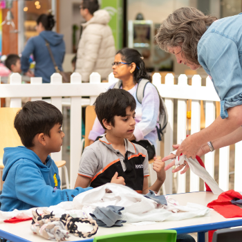 Two young boys at a table learn from an adult showing them how to reuse fabric, in a busy public setting.